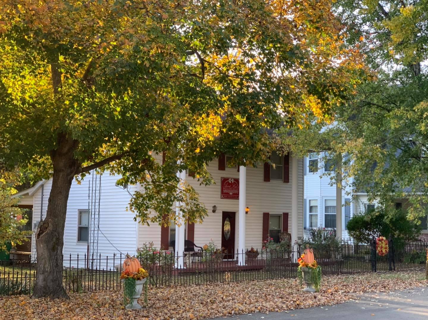 A white two story home with 4 pillars is surrounded by a small lawn covered in fallen autumn leaves. A black cast iron fence implies an established, secure residence. Many yellow leaves remain on trees in front yard.
